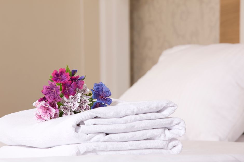 A neatly made bed with white linens and plush pillows in a bedroom featuring a wooden headboard, situated next to a small round bedside table with a marble top. On the table, there is a white ceramic
