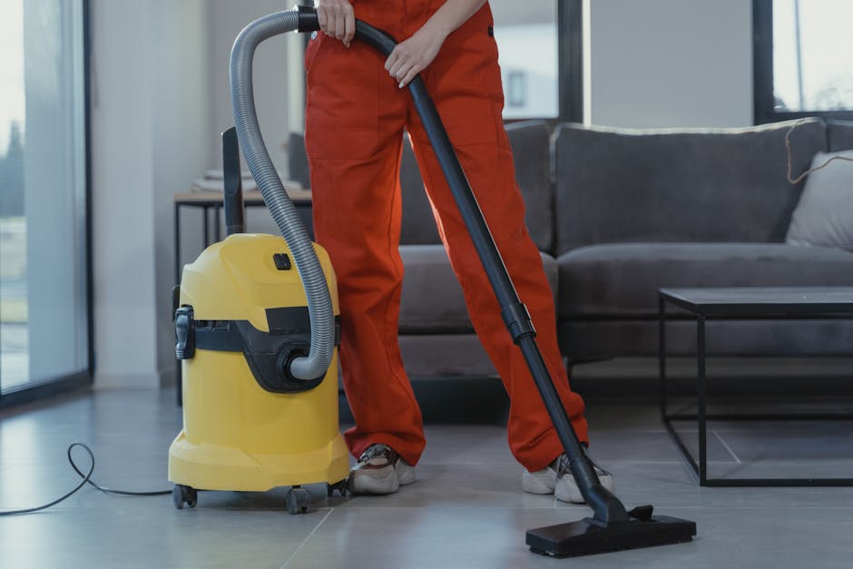 A professional cleaner dressed in white protective suit, mask, and blue boot covers is performing surface cleaning on a beige carpet using a steam cleaning machine. The room features natural light com